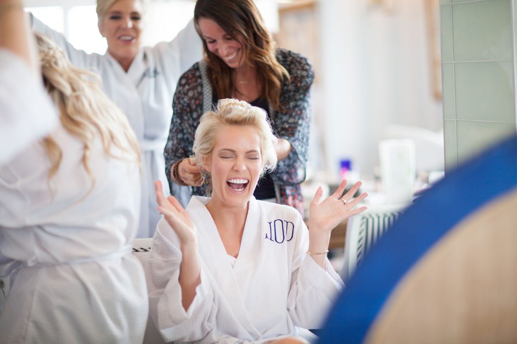 Bride laughing while getting her hair styled in a monogrammed robe before her wedding