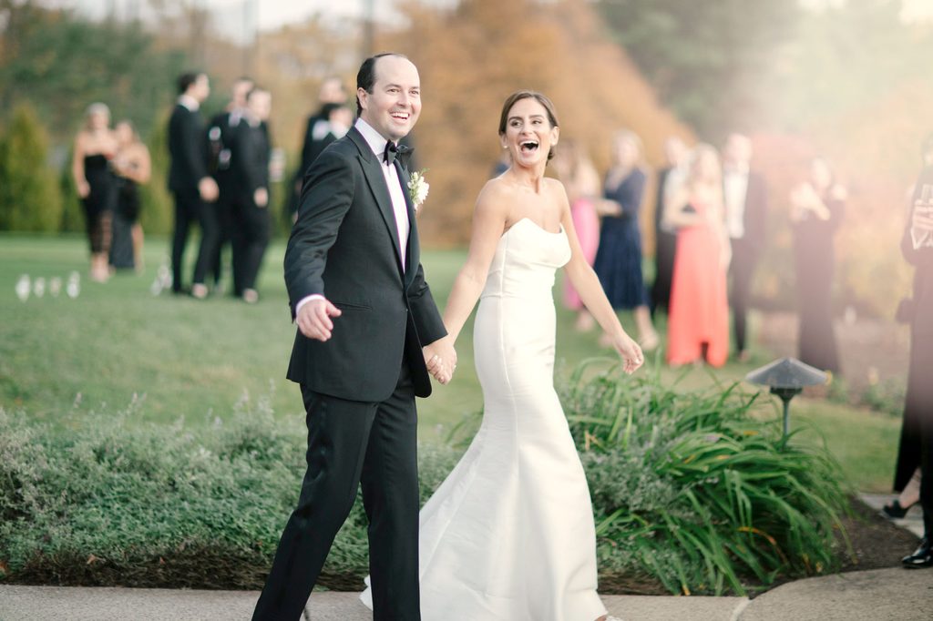 Bride and groom laughing and holding hands as they walk past their wedding guests at golden hour