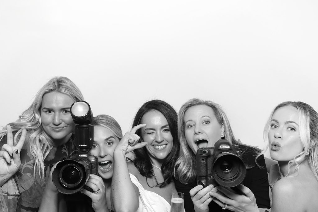 Sarah DiCicco laughing with a group of her brides in a black-and-white photo booth picture, some holding cameras